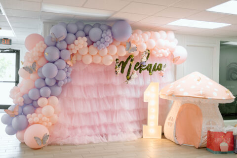 Baby's first birthday photo station with pink ruffles, peach and purple balloons with a lighted "1" and blowup mushroom. Event Planned by Cultivate Event Planning in Oklahoma City.